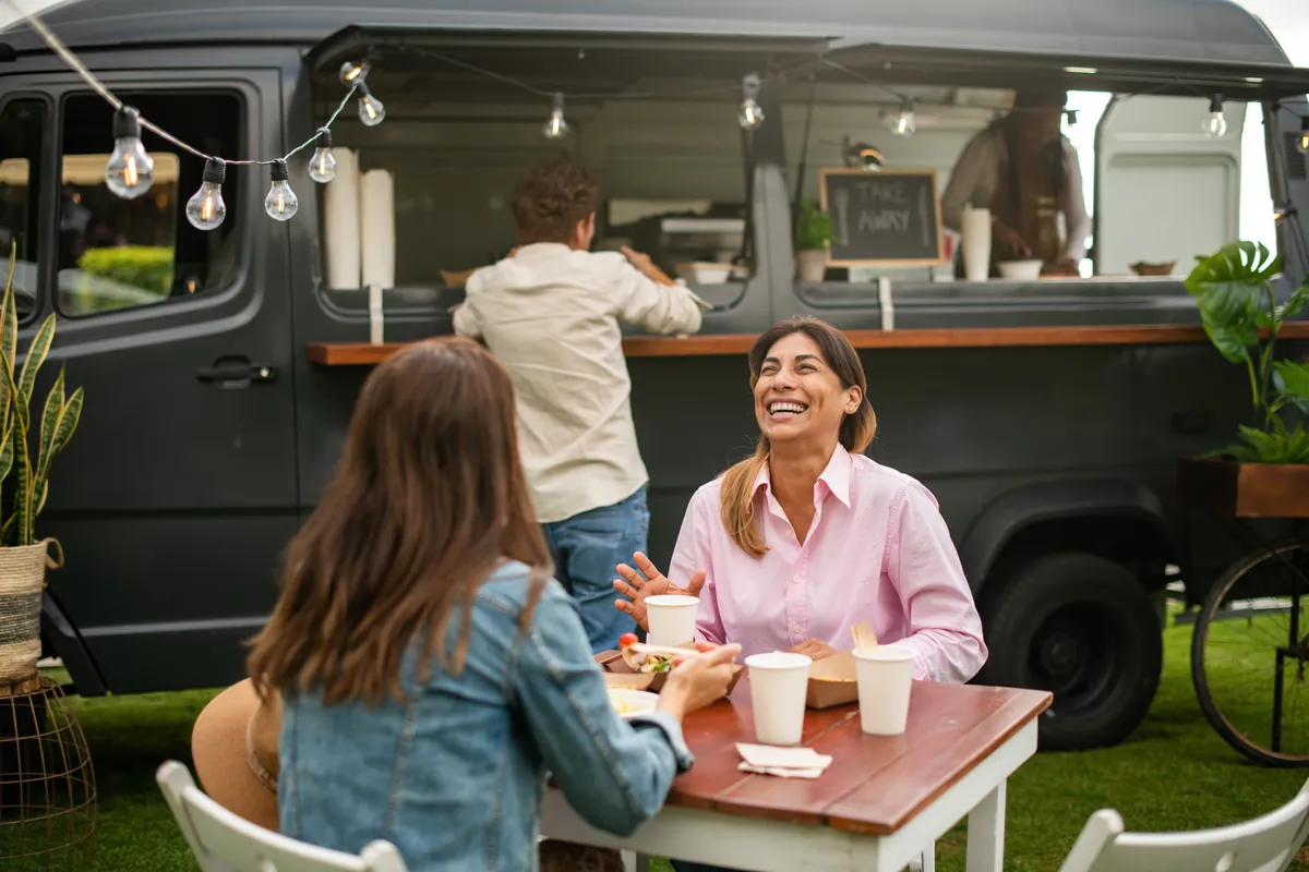 Two women laughing while eating at a table in front of a dark food truck