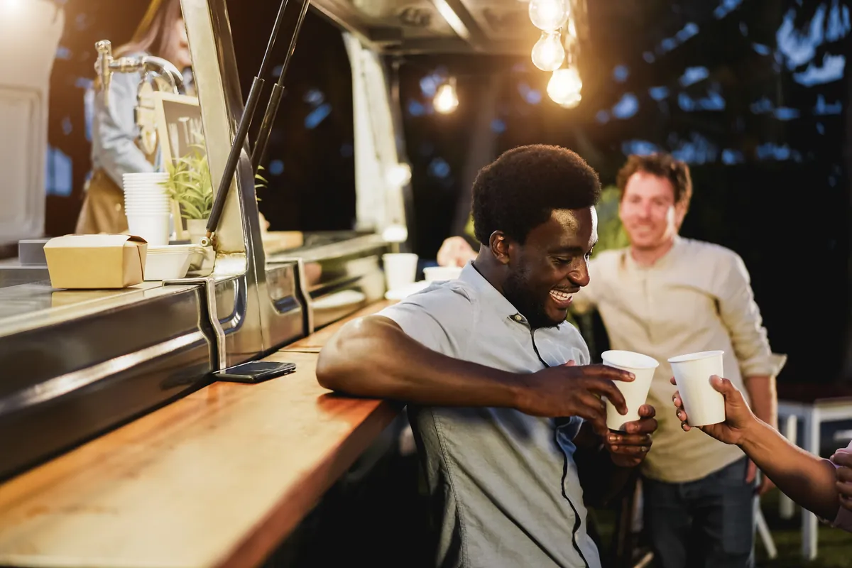 Two guests toasting with cups at a food truck counter in the evening