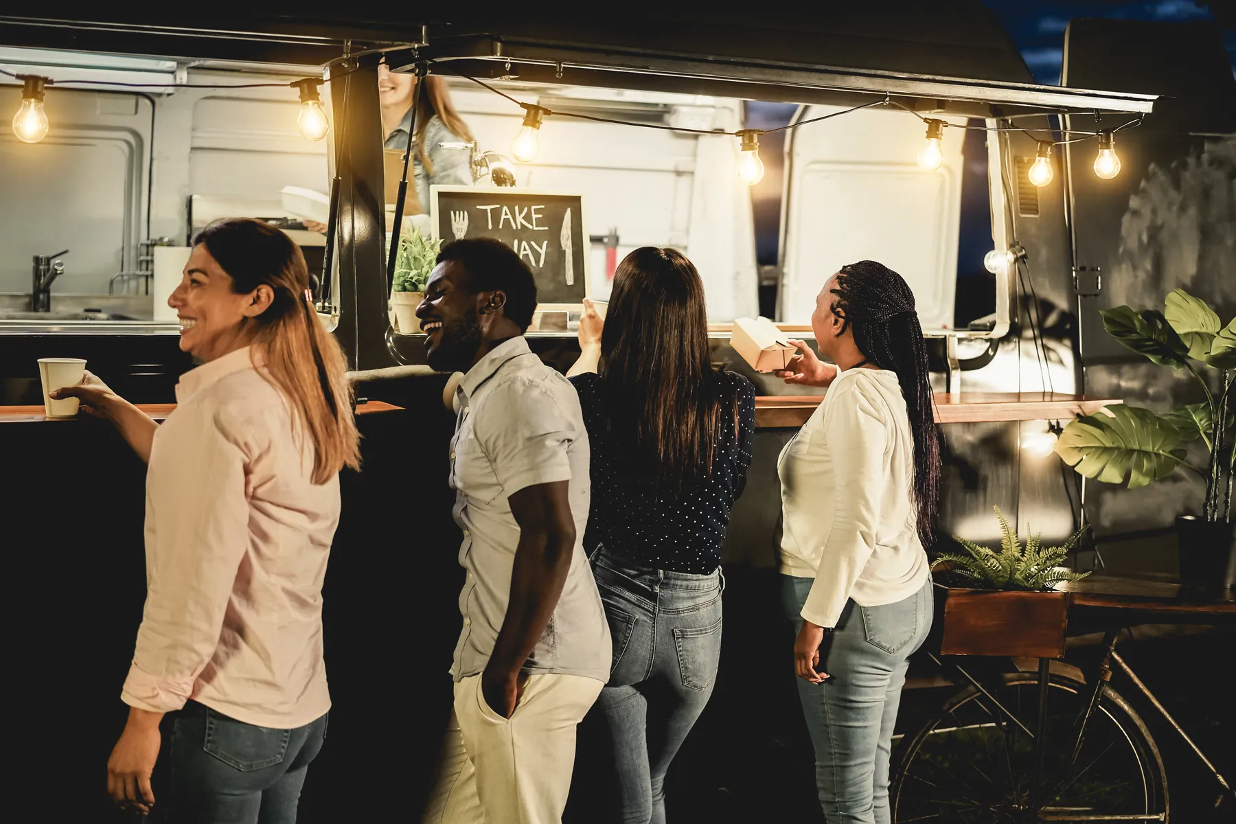 Guests picking up their food in the evening at the window of a food truck with a take-away sign